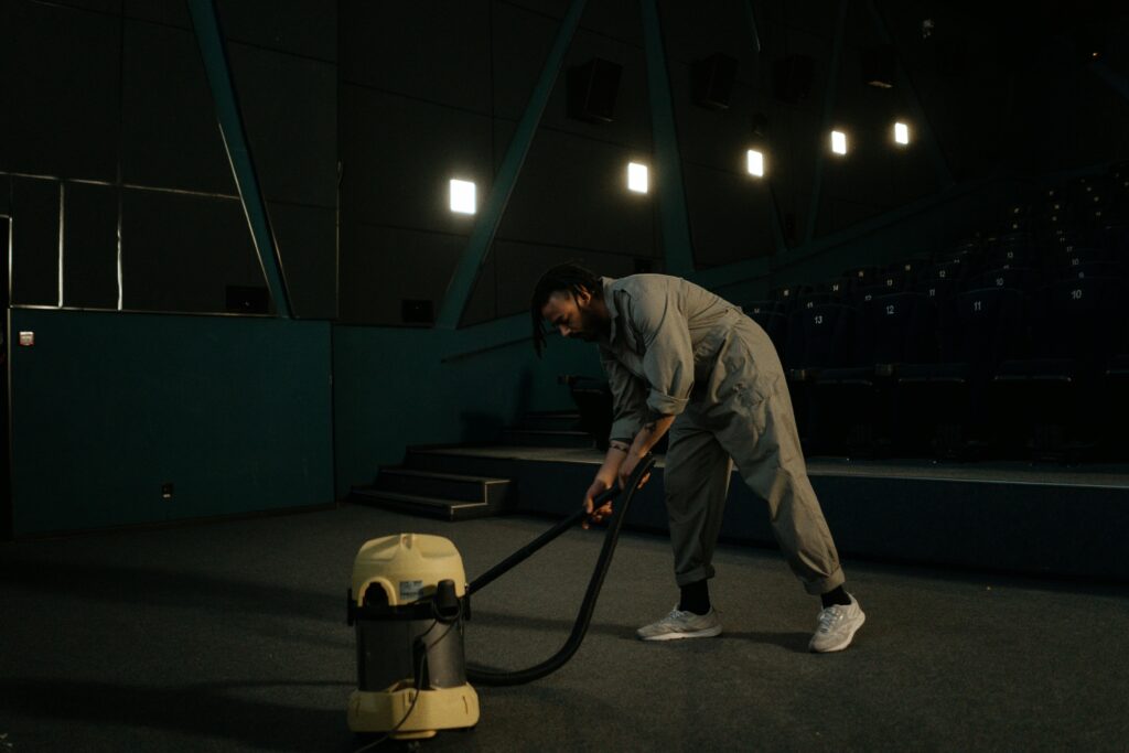 A cinema employee cleaning the theater floor with a vacuum cleaner under dim lights.